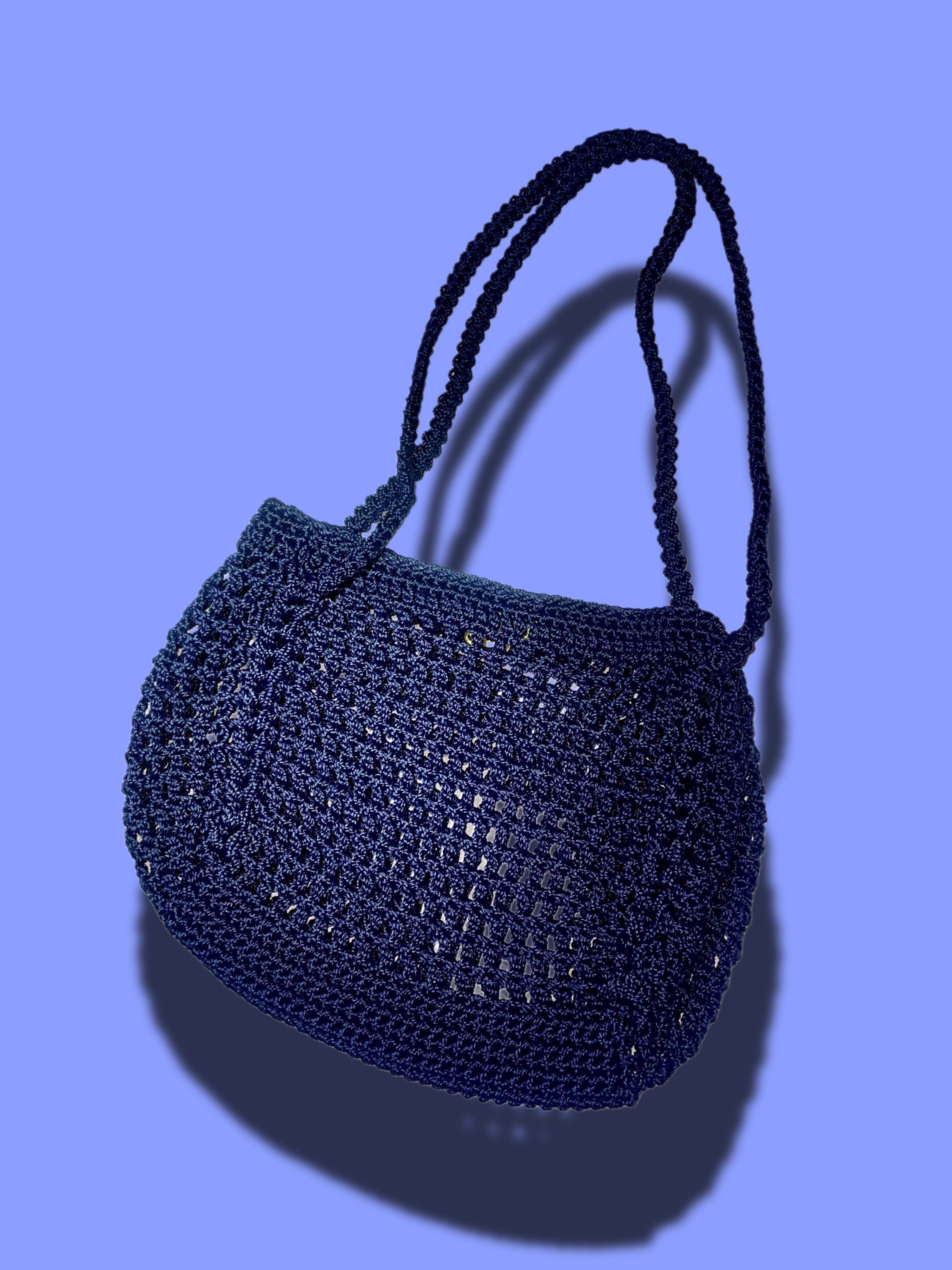 A dark navy blue crocheted shoulder bag with braided shoulder straps, shown against a solid periwinkle blue background with a soft drop shadow.