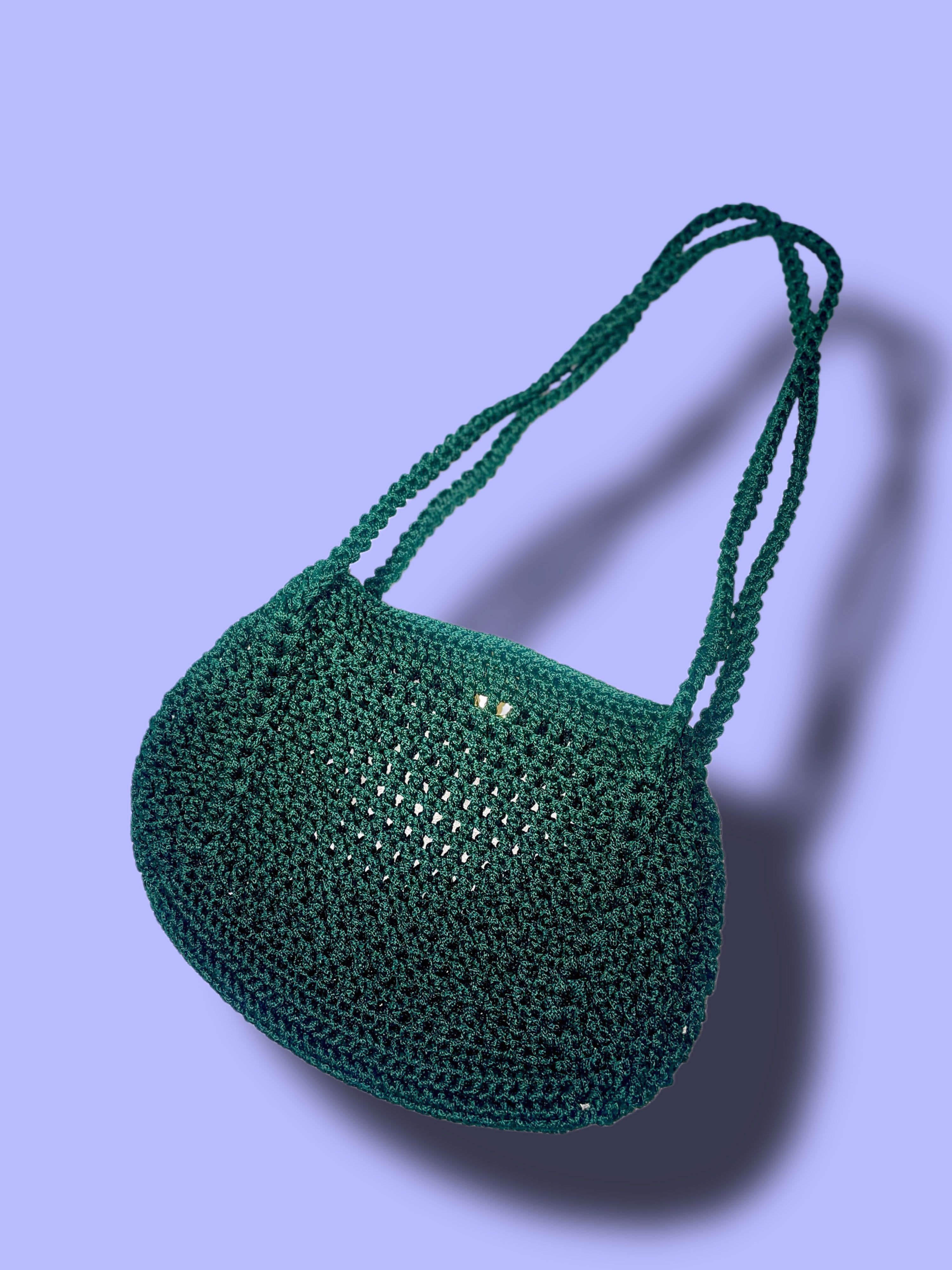 A dark green crocheted shoulder bag with a slouchy, rounded shape and double shoulder straps, shown against a solid lavender background with a soft shadow.
