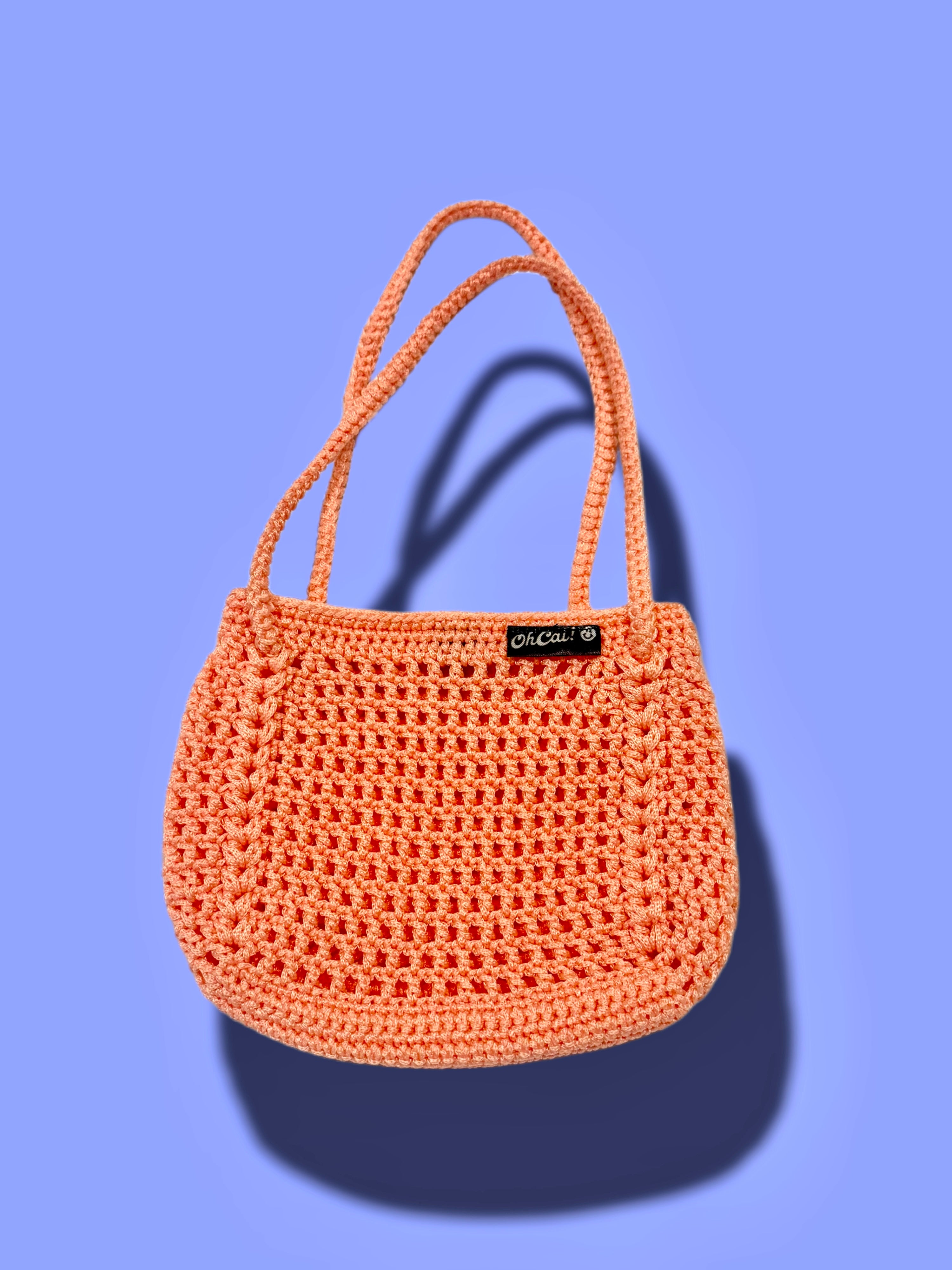 A peach-orange crocheted handbag with two top handles and a small black brand tag reading 'CROCHET' on a solid periwinkle blue background.